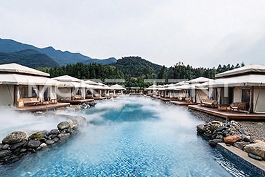 Glamping tents lining a steaming hot spring pool beneath green mountains